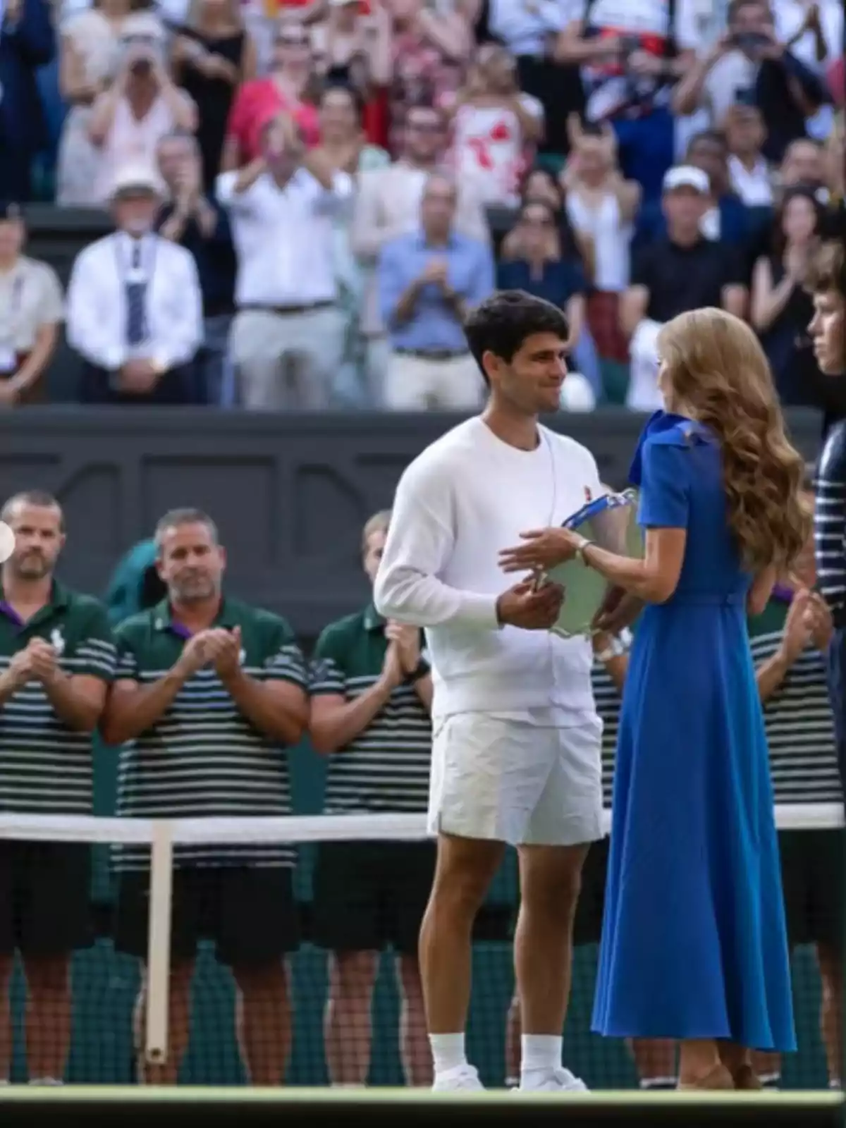 Un tenista recibe un trofeo de una mujer vestida de azul en una cancha de tenis mientras el público y el equipo aplauden al fondo Un tenista recibe un trofeo de una mujer vestida de azul en una cancha de tenis mientras el público y el equipo aplauden al fondo