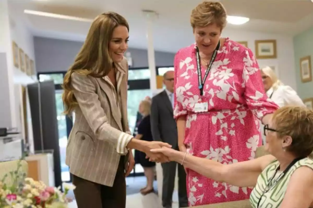 A woman with long, loose hair shakes hands with a seated lady while another woman in a pink dress watches them with a smile in a brightly lit room.