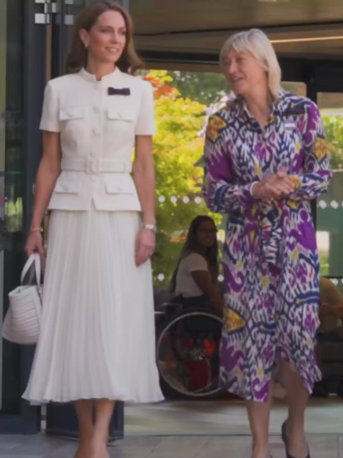 Two women walk together and talk as they leave a building—one is wearing an elegant white dress and the other a colorful dress with a geometric pattern. In the background, a person in a wheelchair can be seen. Two women walk together and talk as they leave a building—one is wearing an elegant white dress and the other a colorful dress with a geometric pattern. In the background, a person in a wheelchair can be seen.