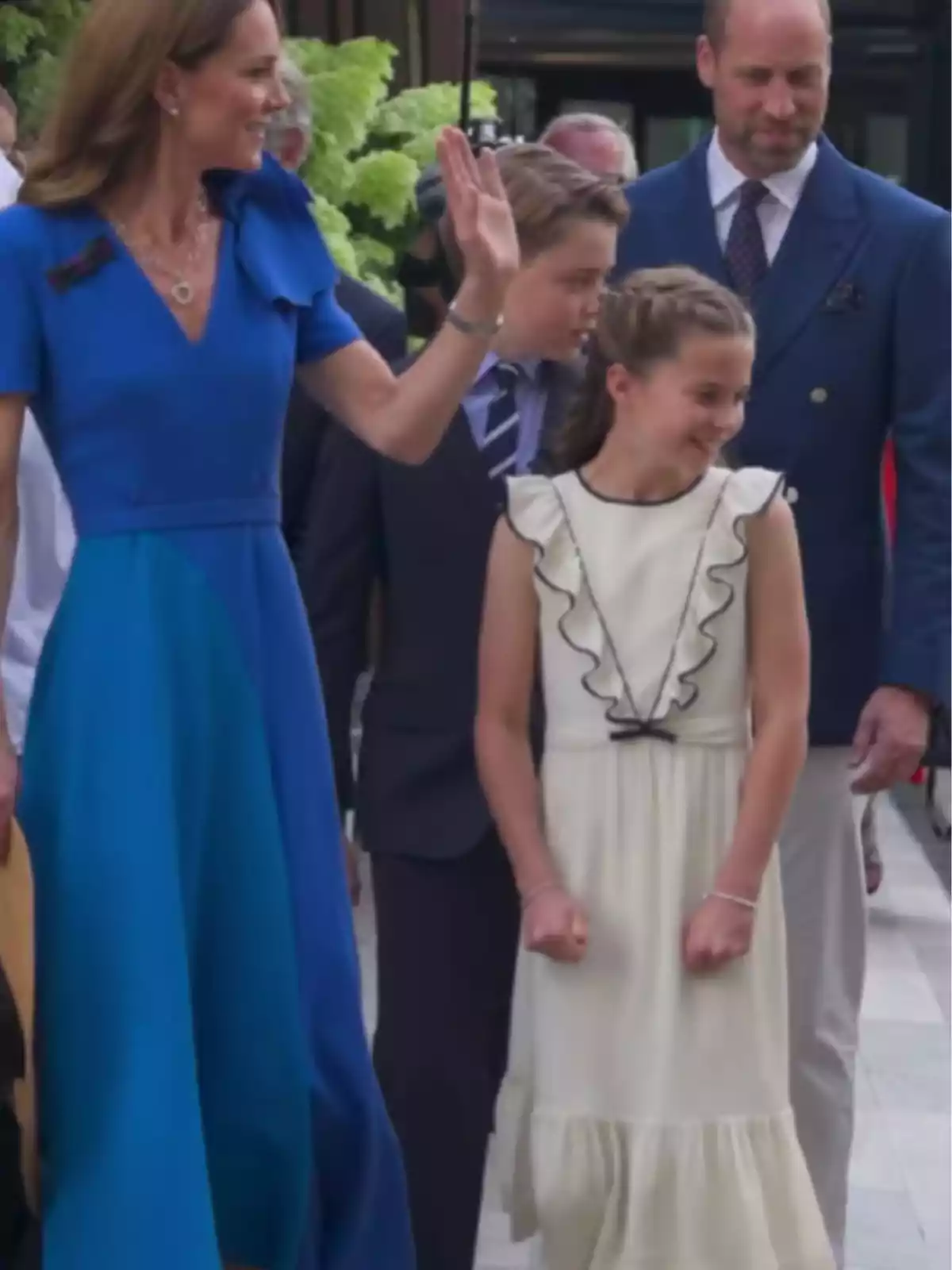 A woman in a blue dress waves while accompanied by a man in a blue jacket, a boy in a suit, and a girl in a white dress; they are all smiling and seem to be at an outdoor event.