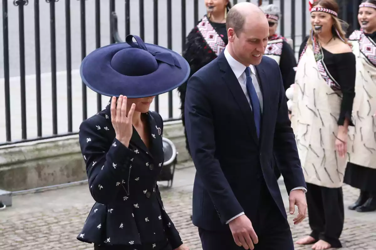 Kate Middleton con un gran sombrero azul camina junto a el príncipe Guillermo con traje oscuro mientras varias personas con vestimenta tradicional sonríen al fondo Kate Middleton con un gran sombrero azul camina junto a el príncipe Guillermo con traje oscuro mientras varias personas con vestimenta tradicional sonríen al fondo