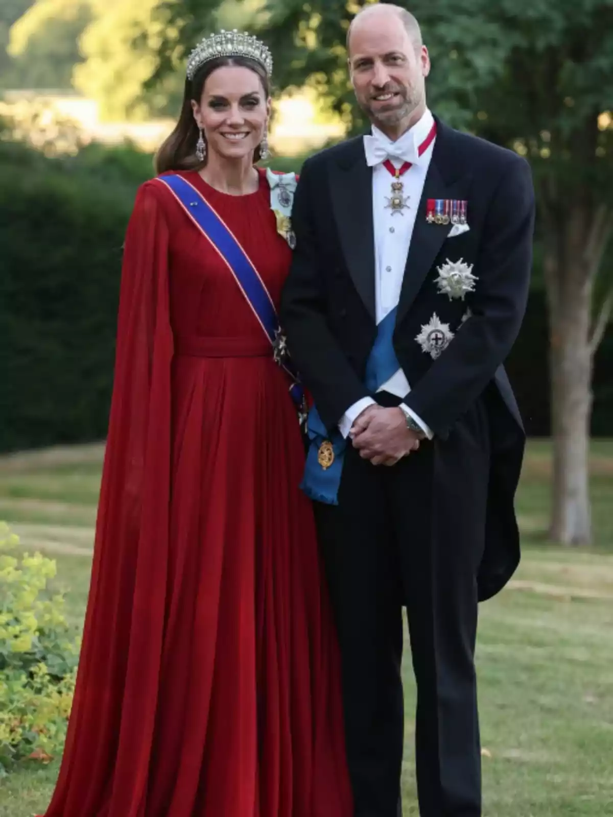 A woman in a long red dress and tiara stands next to a man in a tuxedo with medals as they pose outdoors in a garden.