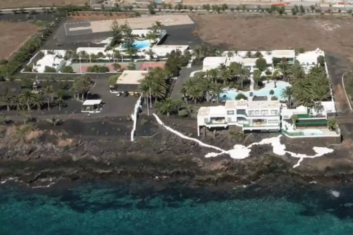 Vista aérea de una gran casa blanca con piscina y jardines ubicada junto al mar con agua turquesa y rodeada de vegetación y terreno árido. Vista aérea de una gran casa blanca con piscina y jardines ubicada junto al mar con agua turquesa y rodeada de vegetación y terreno árido.