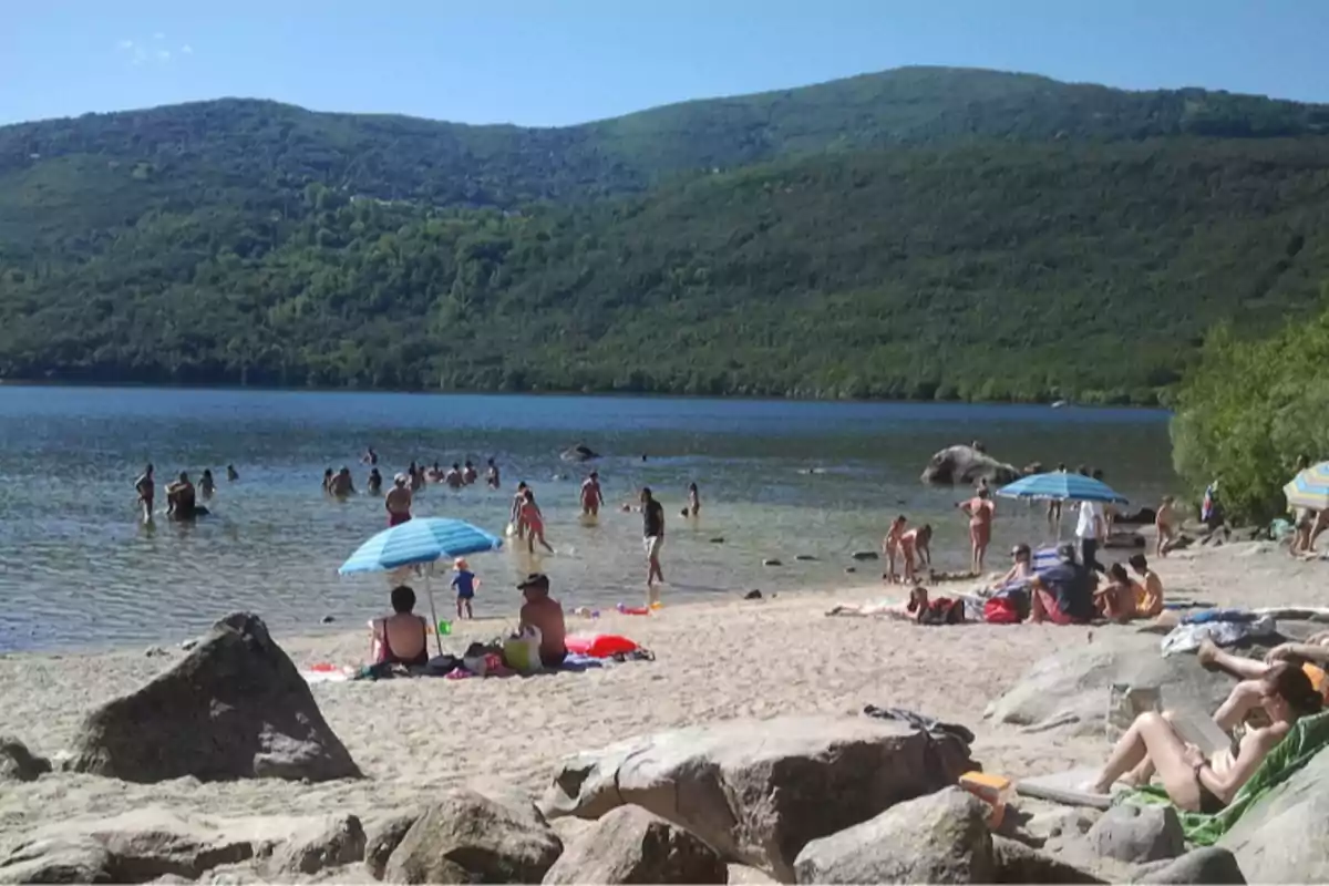 Personas disfrutando de un día soleado en la playa de un lago rodeado de montañas verdes, algunas bajo sombrillas y otras nadando en el agua Personas disfrutando de un día soleado en la playa de un lago rodeado de montañas verdes, algunas bajo sombrillas y otras nadando en el agua