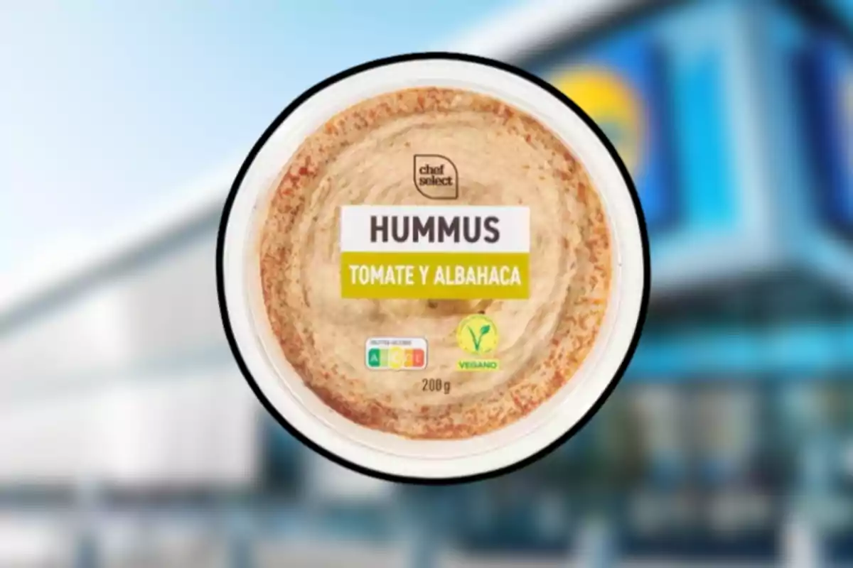 Envase de hummus sabor tomate y albahaca de la marca Chef Select con etiqueta vegana sobre un fondo desenfocado de supermercado. Envase de hummus sabor tomate y albahaca de la marca Chef Select con etiqueta vegana sobre un fondo desenfocado de supermercado.
