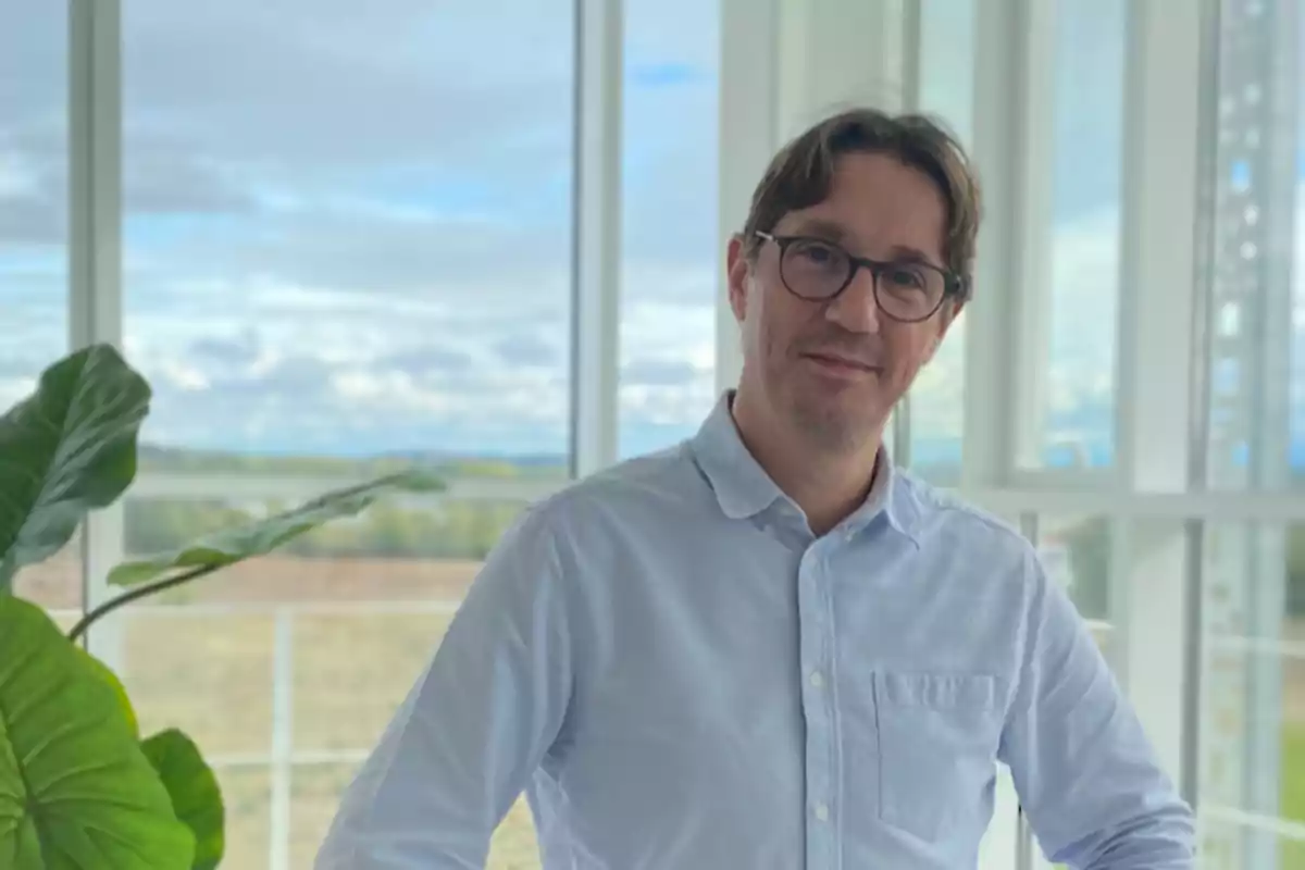 Hombre con gafas y camisa azul claro posando junto a una ventana grande con vista al exterior y una planta verde a su lado