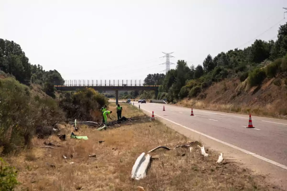 Trabajadores con chalecos reflectantes revisan restos de un accidente en la cuneta de una carretera junto a conos naranjas y un puente al fondo Trabajadores con chalecos reflectantes revisan restos de un accidente en la cuneta de una carretera junto a conos naranjas y un puente al fondo
