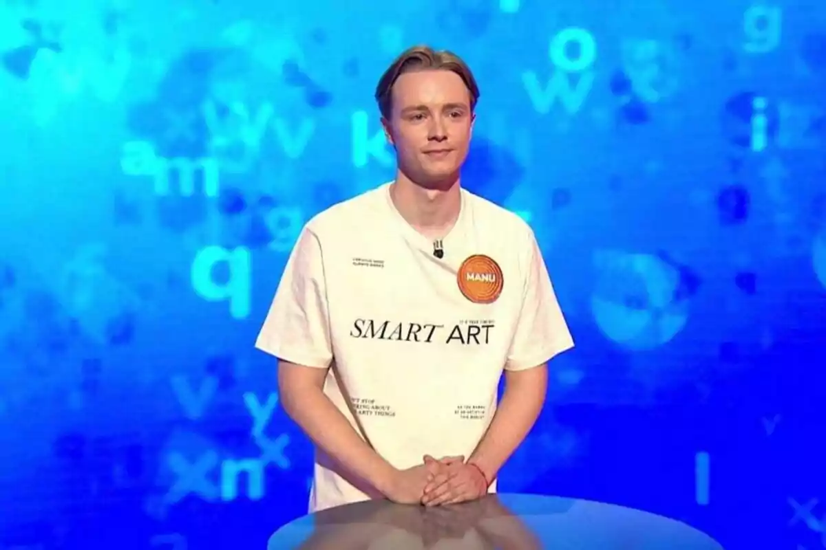 Manu Pascual, de pie detrás de un atril en un set de televisión con fondo azul y letras dispersas, lleva una camiseta blanca con el texto SMART ART y un círculo naranja que dice MANU. Manu Pascual, de pie detrás de un atril en un set de televisión con fondo azul y letras dispersas, lleva una camiseta blanca con el texto SMART ART y un círculo naranja que dice MANU.