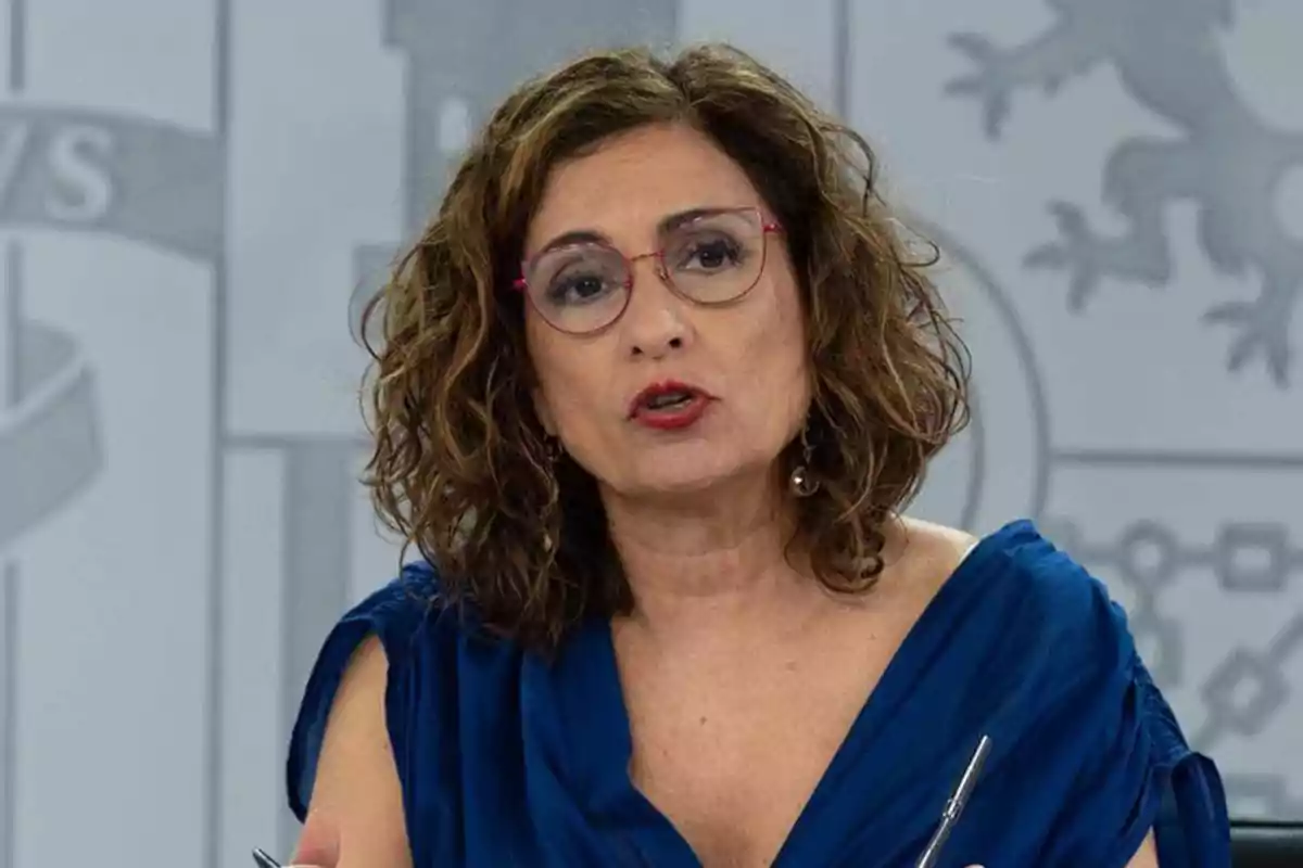 Mujer de cabello rizado y gafas con montura rosa hablando frente a un micrófono y usando un vestido azul Mujer de cabello rizado y gafas con montura rosa hablando frente a un micrófono y usando un vestido azul