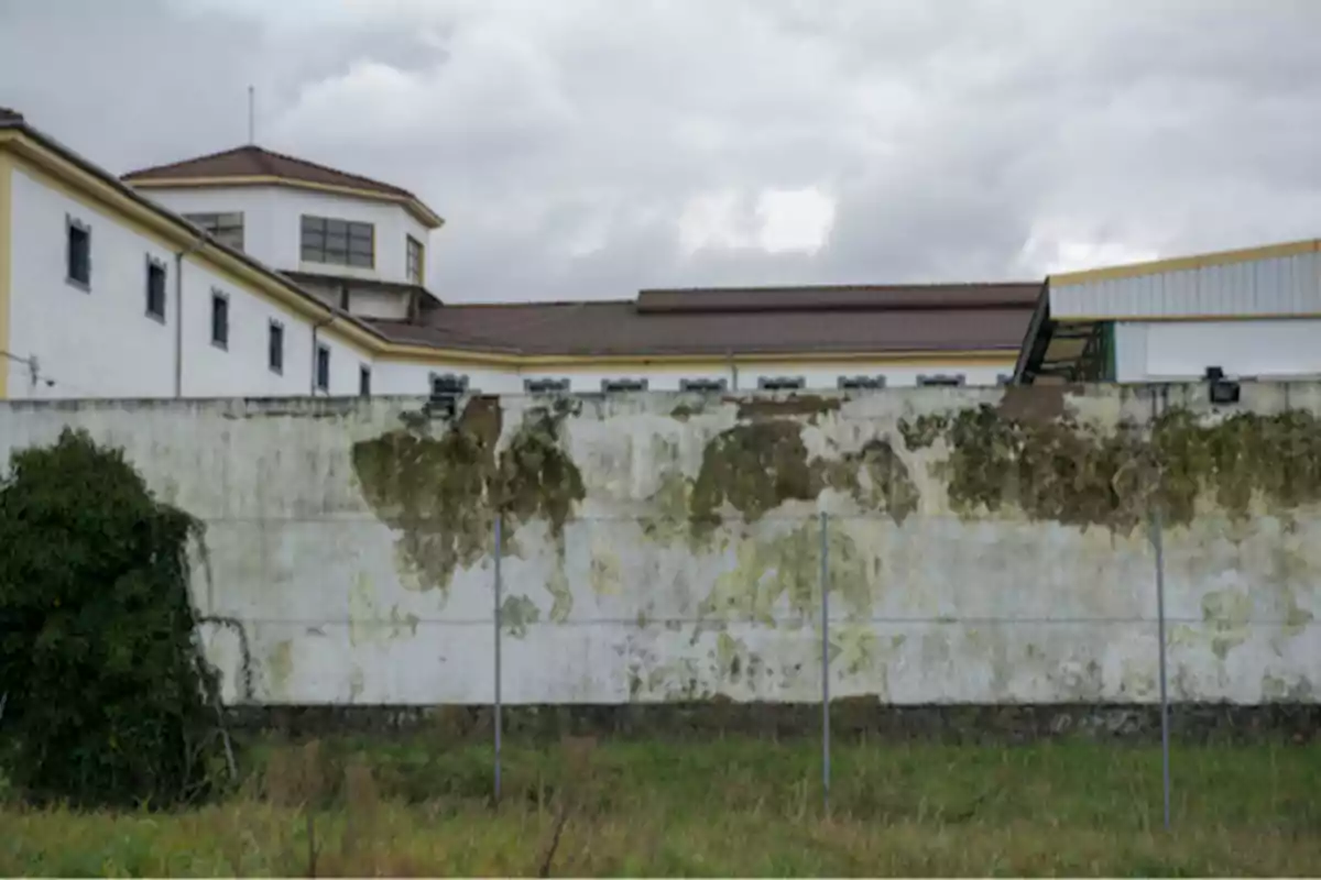 Edificio de paredes blancas con manchas de humedad y una cerca metálica al frente bajo un cielo nublado Edificio de paredes blancas con manchas de humedad y una cerca metálica al frente bajo un cielo nublado