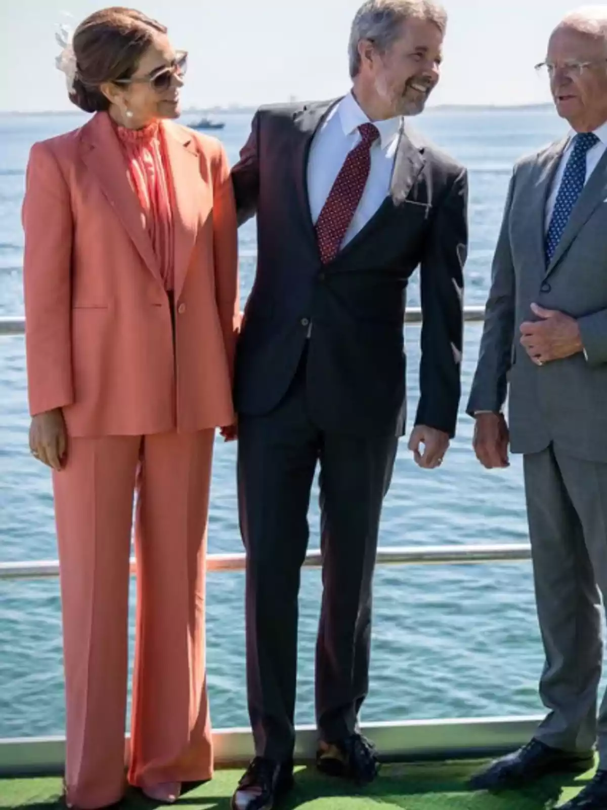 Tres personas elegantemente vestidas conversan y sonríen de pie sobre una plataforma junto al mar en un día soleado. Tres personas elegantemente vestidas conversan y sonríen de pie sobre una plataforma junto al mar en un día soleado.