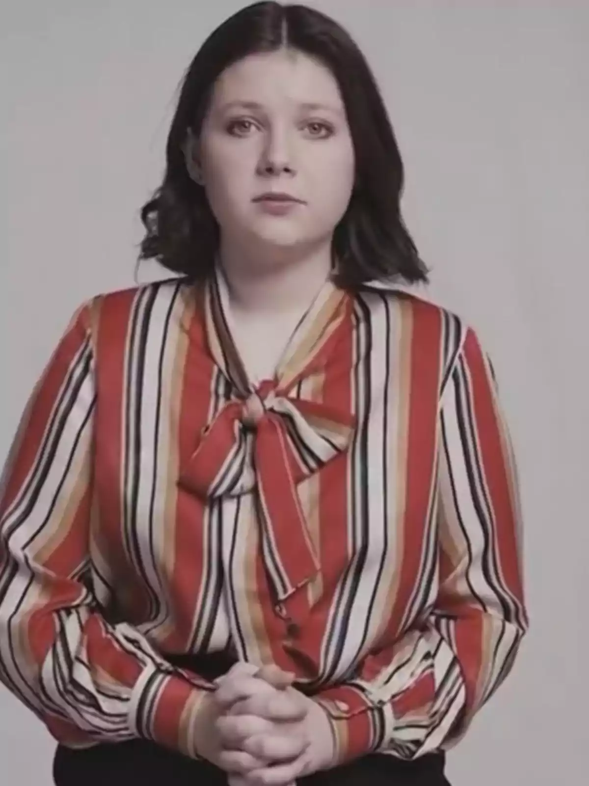 Dark-haired woman wearing a red, white, and black striped blouse, looking straight ahead with her hands clasped. Dark-haired woman wearing a red, white, and black striped blouse, looking straight ahead with her hands clasped.