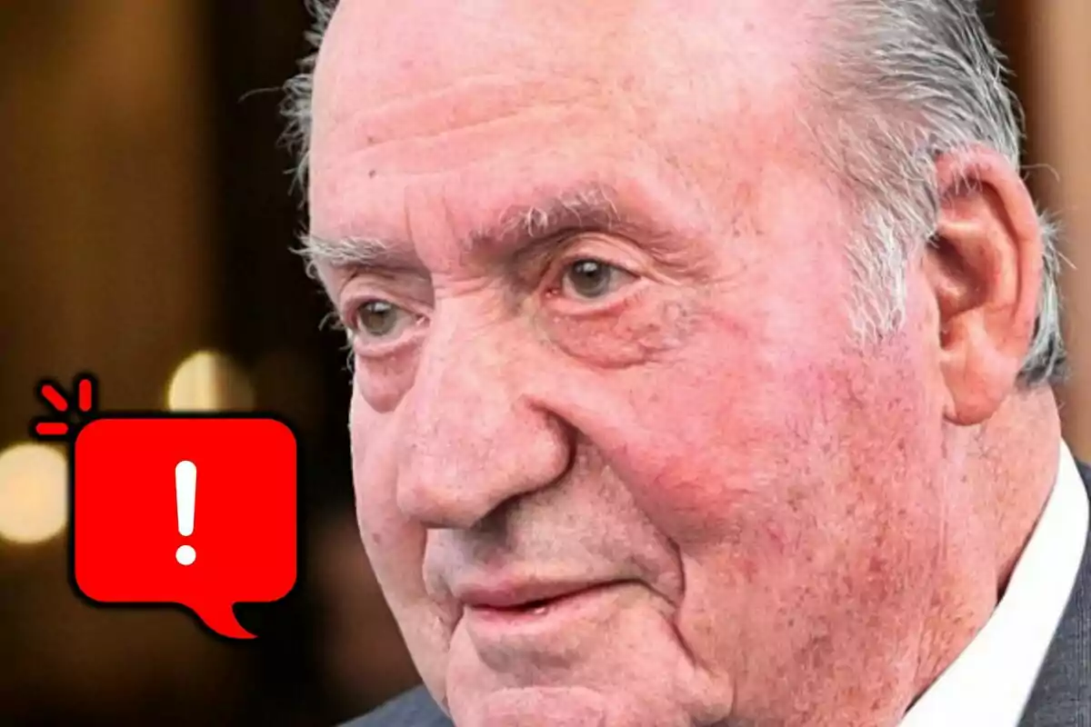 Primer plano de Juan Carlos I con expresión seria y un ícono de advertencia en color rojo a la izquierda.