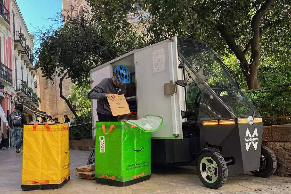Un repartidor con casco azul organiza paquetes de Amazon junto a un vehículo eléctrico de reparto en una calle urbana con árboles y edificios alrededor