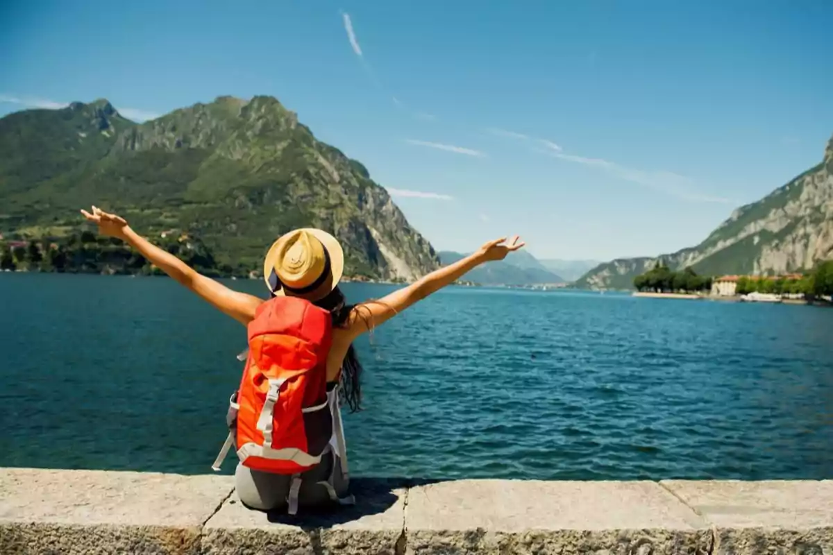 Mujer con sombrero y mochila roja sentada frente a un lago rodeado de montañas con los brazos extendidos disfrutando del paisaje