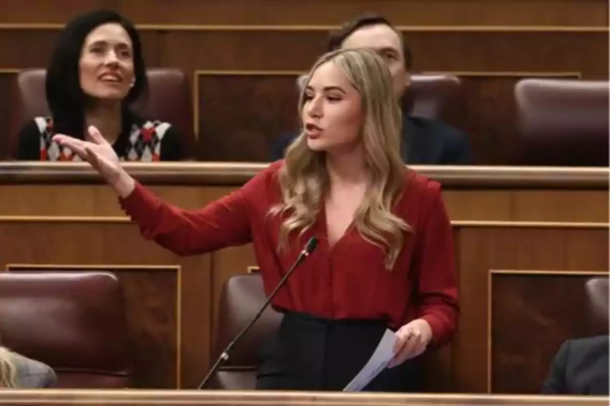 Mujer joven de cabello rubio y suelto hablando en un parlamento mientras sostiene unos papeles y gesticula con la mano derecha Mujer joven de cabello rubio y suelto hablando en un parlamento mientras sostiene unos papeles y gesticula con la mano derecha