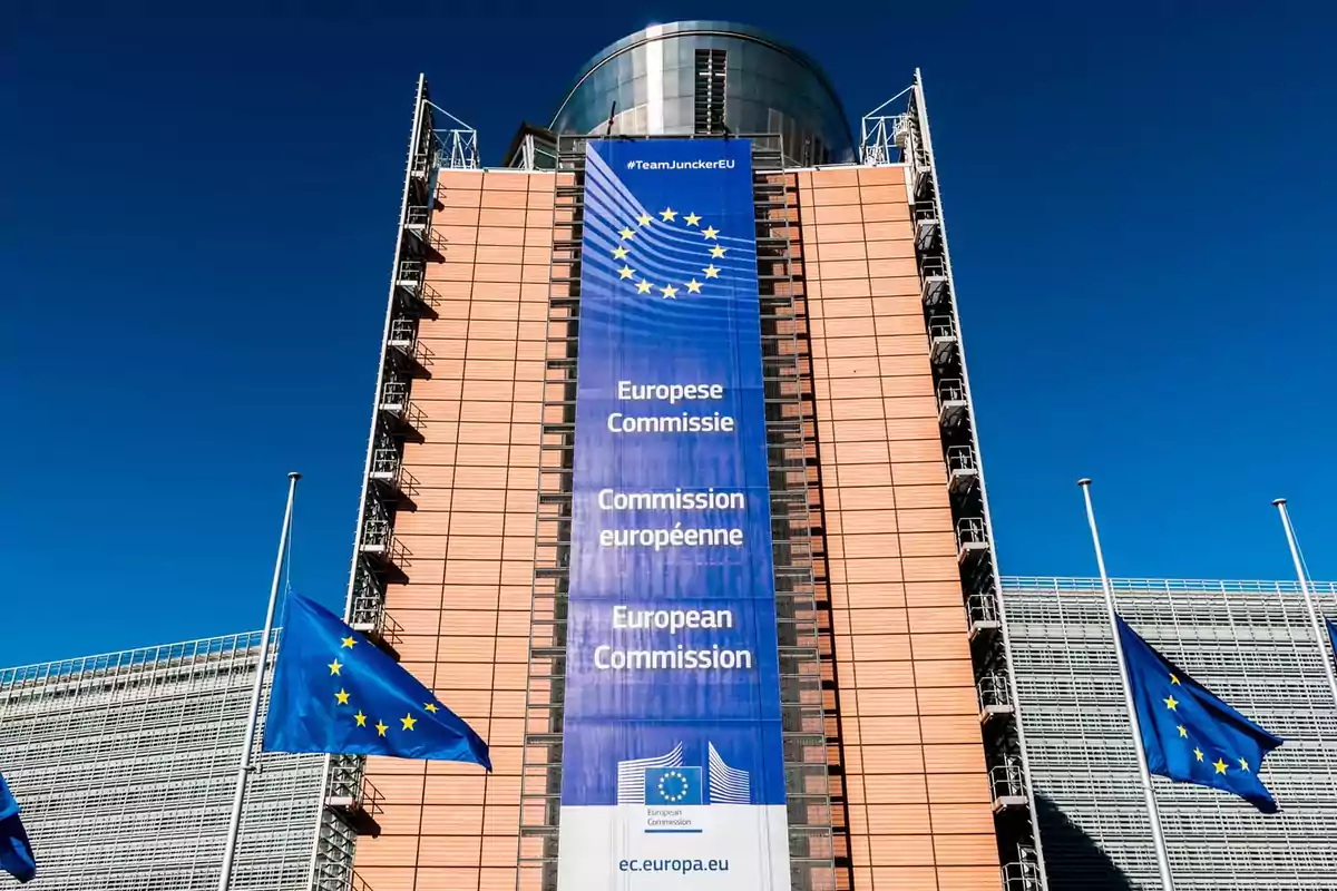 Edificio de la Comisión Europea con una gran pancarta azul y banderas de la Unión Europea ondeando frente a un cielo despejado