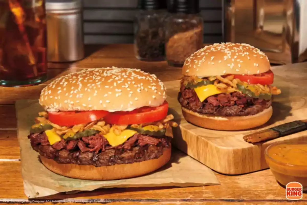 Dos hamburguesas con pan de ajonjolí, carne, queso, jitomate, pepinillos y cebolla crujiente sobre una mesa de madera junto a un vaso de bebida y un recipiente pequeño de salsa Dos hamburguesas con pan de ajonjolí, carne, queso, jitomate, pepinillos y cebolla crujiente sobre una mesa de madera junto a un vaso de bebida y un recipiente pequeño de salsa