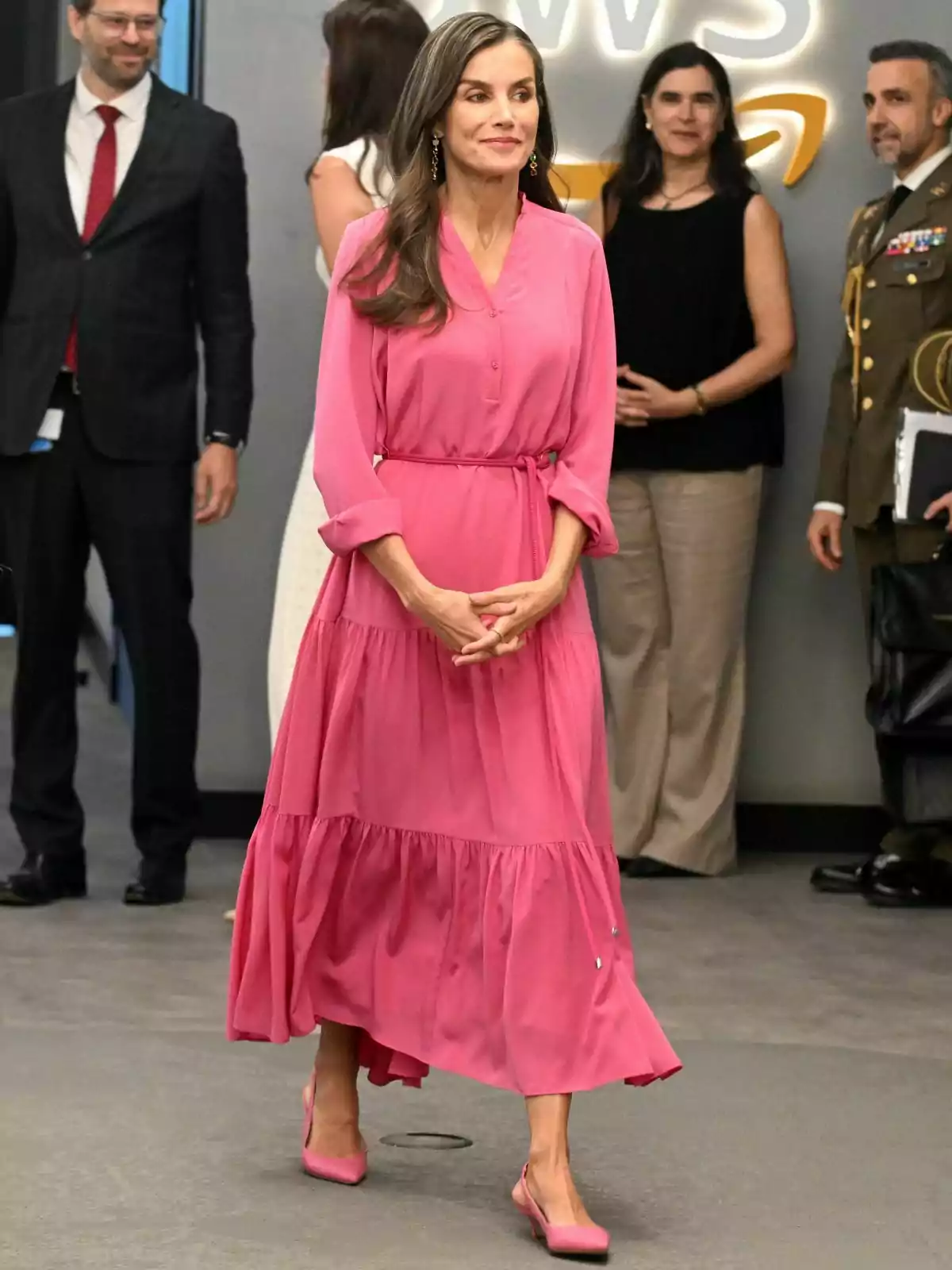 La reina Letizia Ortiz con vestido largo rosa y zapatos a juego caminando en un evento formal con varias personas al fondo. La reina Letizia Ortiz con vestido largo rosa y zapatos a juego caminando en un evento formal con varias personas al fondo.