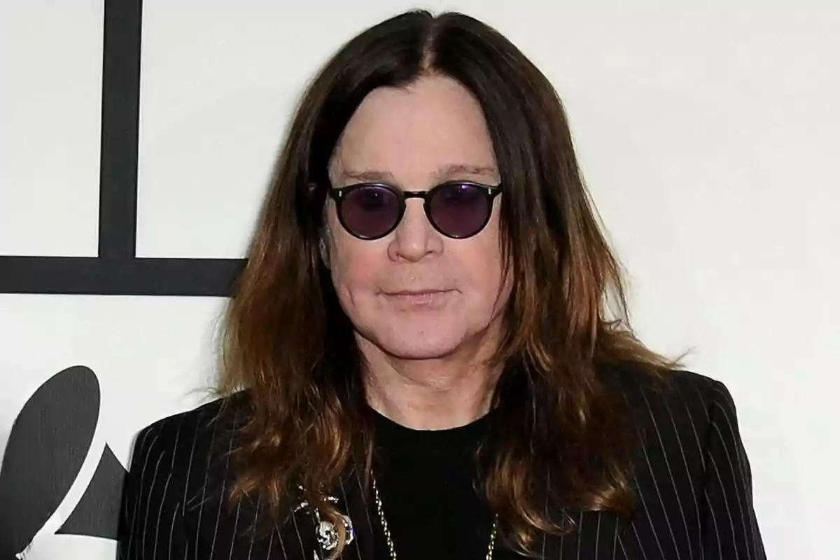 Ozzy Osbourne with long brown hair, round sunglasses, and a striped jacket posing in front of a light background