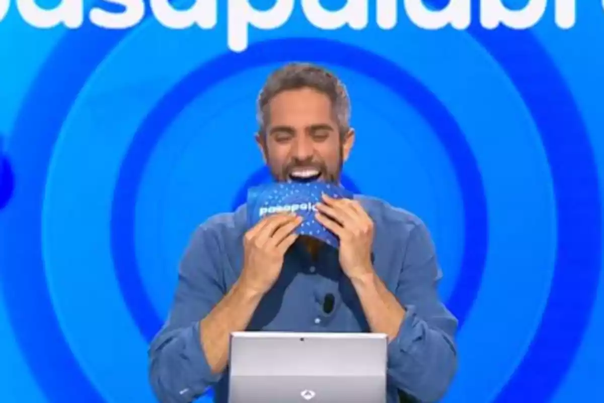 Roberto Leal sonriente con barba y camisa azul sostiene una tarjeta azul de Pasapalabra frente a su boca en un set de televisión con fondo azul Roberto Leal sonriente con barba y camisa azul sostiene una tarjeta azul de Pasapalabra frente a su boca en un set de televisión con fondo azul