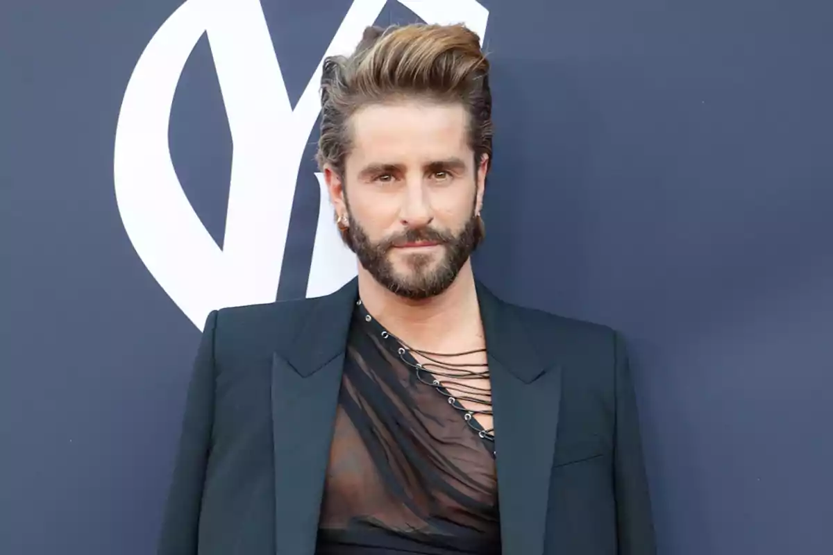 Pelayo Díaz usando un saco negro y una camiseta transparente con detalles de cordones posando frente a un fondo azul con un logotipo blanco. Pelayo Díaz usando un saco negro y una camiseta transparente con detalles de cordones posando frente a un fondo azul con un logotipo blanco.