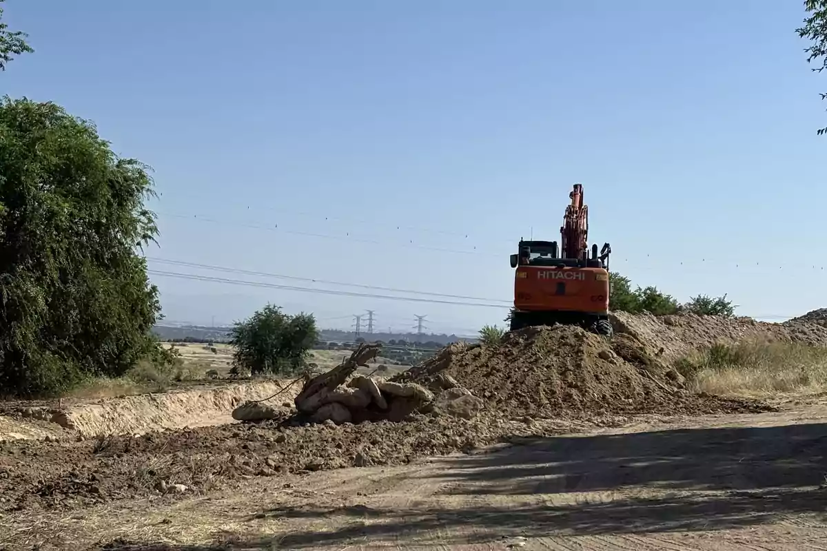 Excavadora naranja sobre un montículo de tierra en una zona rural con árboles y líneas eléctricas al fondo Excavadora naranja sobre un montículo de tierra en una zona rural con árboles y líneas eléctricas al fondo