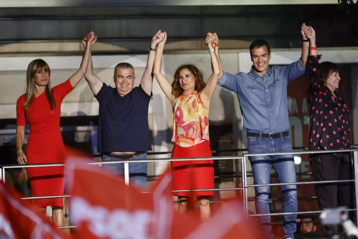 Cinco personas de pie en un escenario al aire libre levantan las manos unidas mientras sonríen y al frente se ven banderas rojas ondeando entre el público en Ferraz
