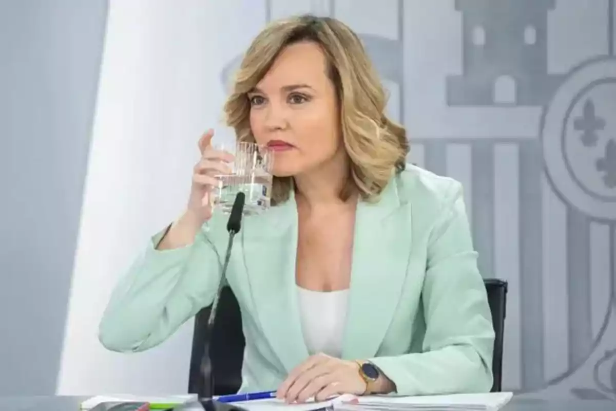 Mujer rubia con saco verde claro sentada en una mesa tomando un vaso de agua frente a un micrófono Mujer rubia con saco verde claro sentada en una mesa tomando un vaso de agua frente a un micrófono