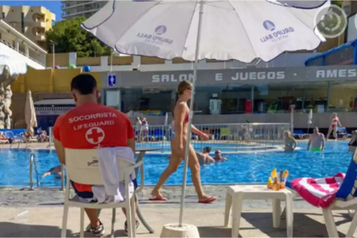 Un socorrista sentado bajo una sombrilla observa una piscina donde varias personas disfrutan del agua y el sol Un socorrista sentado bajo una sombrilla observa una piscina donde varias personas disfrutan del agua y el sol