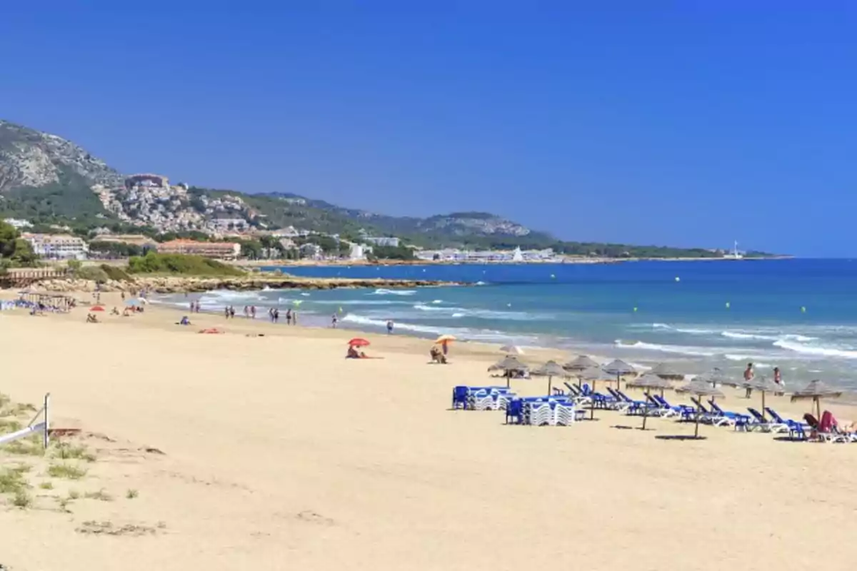 Playa de arena dorada con sombrillas y tumbonas, personas disfrutando del sol y el mar, montañas y edificios al fondo bajo un cielo despejado Playa de arena dorada con sombrillas y tumbonas, personas disfrutando del sol y el mar, montañas y edificios al fondo bajo un cielo despejado