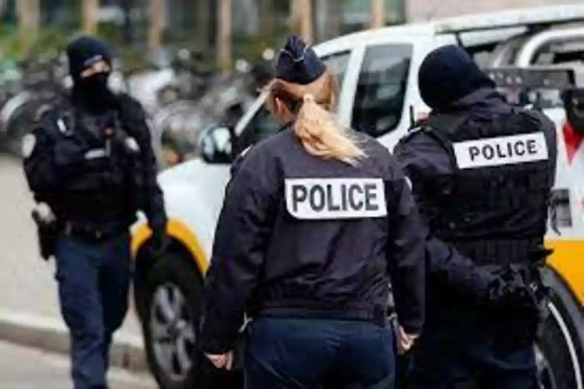 Tres agentes de policía con uniformes oscuros están de pie junto a un vehículo policial en la calle. Tres agentes de policía con uniformes oscuros están de pie junto a un vehículo policial en la calle.