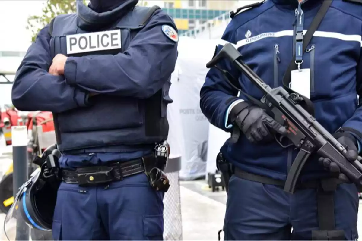Dos agentes de policía con uniforme azul y chaleco antibalas uno con los brazos cruzados y el otro sosteniendo un arma larga
