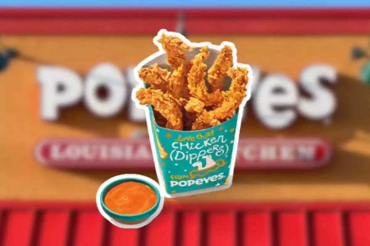Tiras de pollo empanizadas en un recipiente de Popeyes acompañadas de una salsa frente a un fondo borroso del restaurante Tiras de pollo empanizadas en un recipiente de Popeyes acompañadas de una salsa frente a un fondo borroso del restaurante