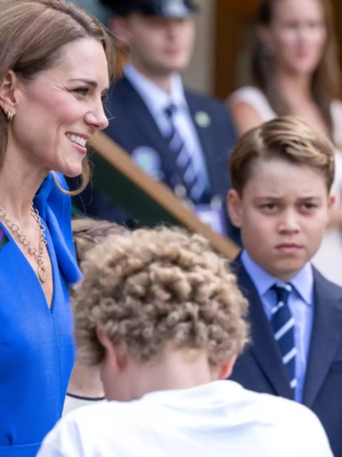 Kate Middleton sonriente con vestido azul está acompañada del príncipe George con traje y corbata y otro niño de cabello rizado en primer plano, mientras varias personas observan al fondo Kate Middleton sonriente con vestido azul está acompañada del príncipe George con traje y corbata y otro niño de cabello rizado en primer plano, mientras varias personas observan al fondo