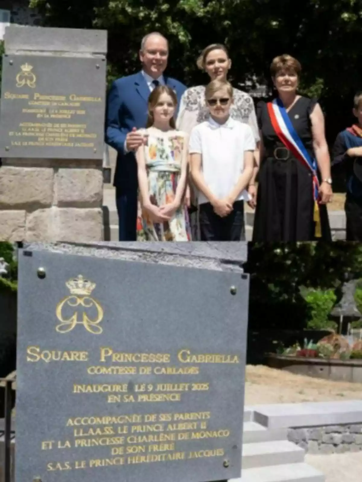 Un grupo de personas posa junto a una placa conmemorativa de piedra gris con letras doradas en un espacio al aire libre rodeado de árboles y jardines Un grupo de personas posa junto a una placa conmemorativa de piedra gris con letras doradas en un espacio al aire libre rodeado de árboles y jardines