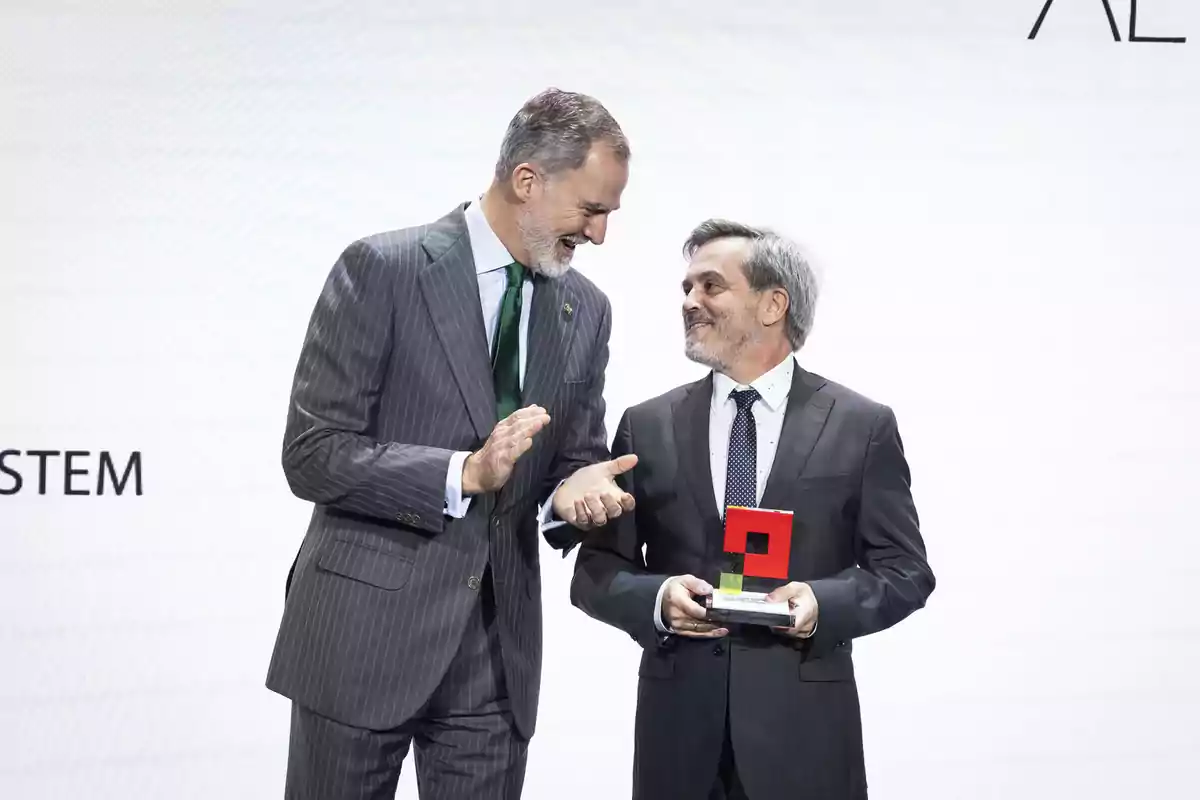 Dos hombres con traje sonríen mientras uno de ellos sostiene un trofeo rojo en una ceremonia de premiación Dos hombres con traje sonríen mientras uno de ellos sostiene un trofeo rojo en una ceremonia de premiación
