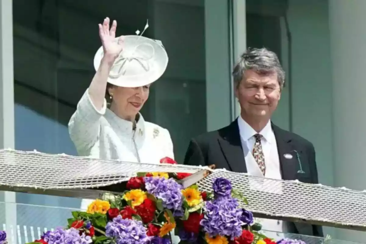 A woman dressed in white with a hat waves while standing next to a man in a dark suit, both behind a railing decorated with colorful flowers. A woman dressed in white with a hat waves while standing next to a man in a dark suit, both behind a railing decorated with colorful flowers.