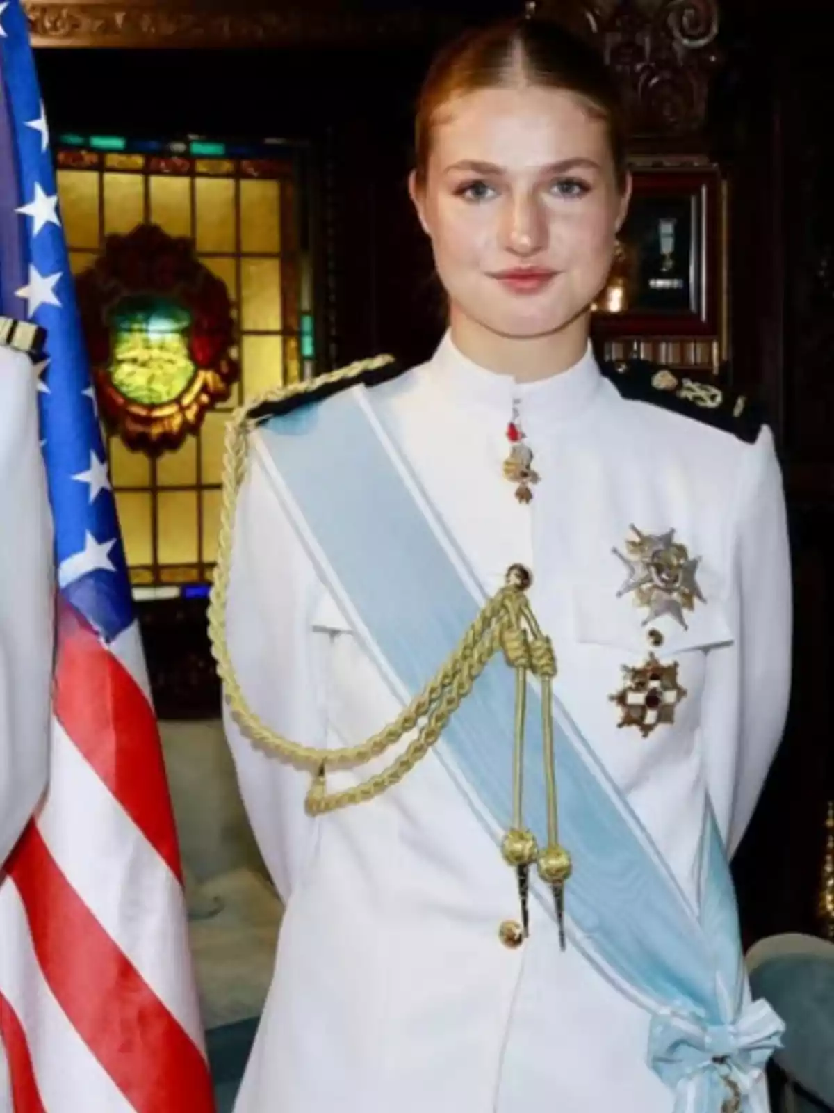 Joven con uniforme militar blanco con charreteras doradas y banda azul claro, de pie junto a una bandera de Estados Unidos en un entorno elegante Joven con uniforme militar blanco con charreteras doradas y banda azul claro, de pie junto a una bandera de Estados Unidos en un entorno elegante