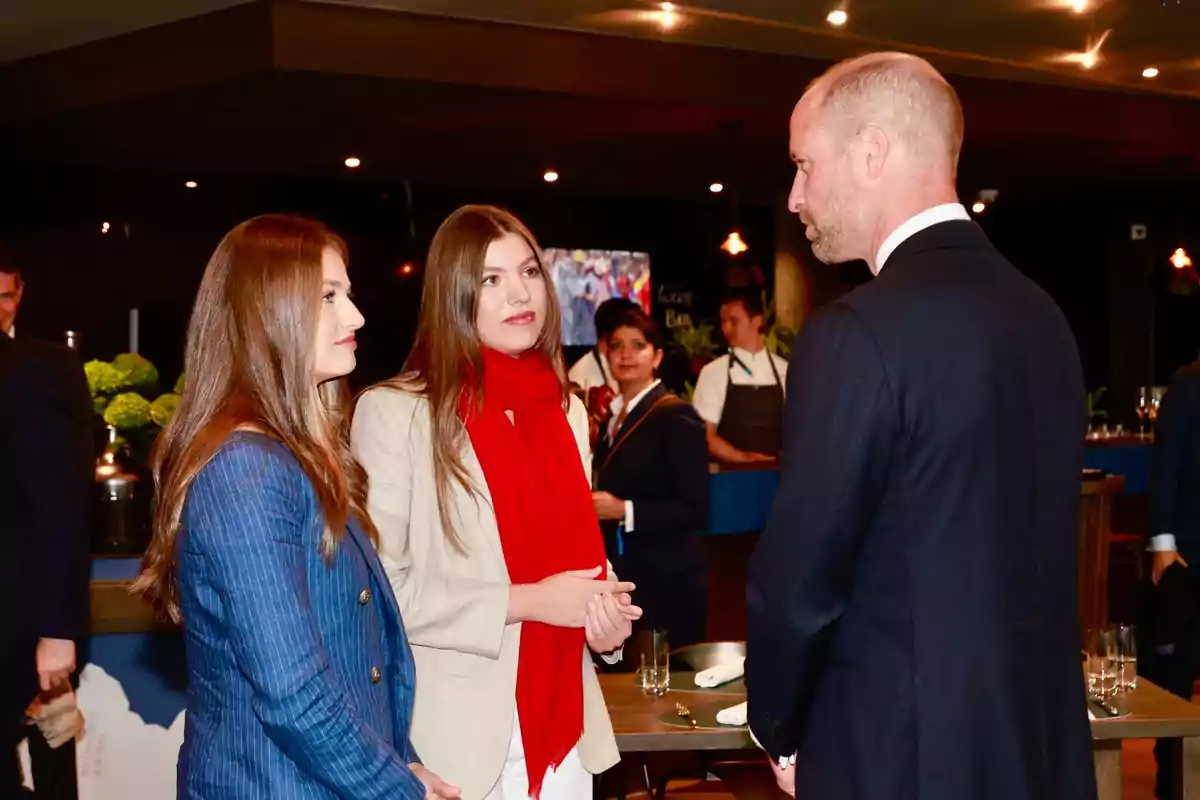 Leonor and Sofía talk with Prince William in an elegant setting with people in the background. Leonor and Sofía talk with Prince William in an elegant setting with people in the background.