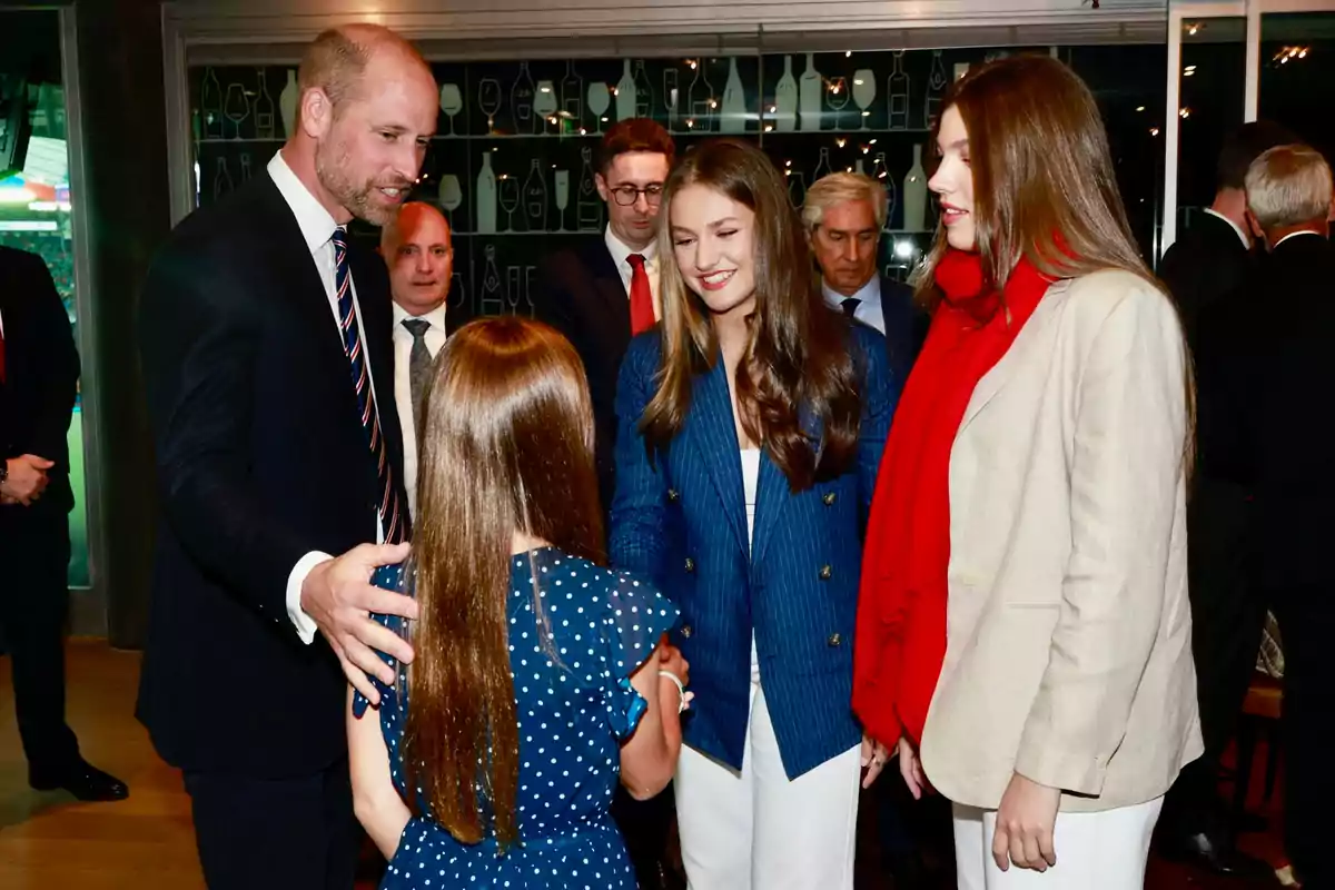 A man in a suit greets a girl while two young people smile and watch the scene in a formal setting with several people in the background. A man in a suit greets a girl while two young people smile and watch the scene in a formal setting with several people in the background.