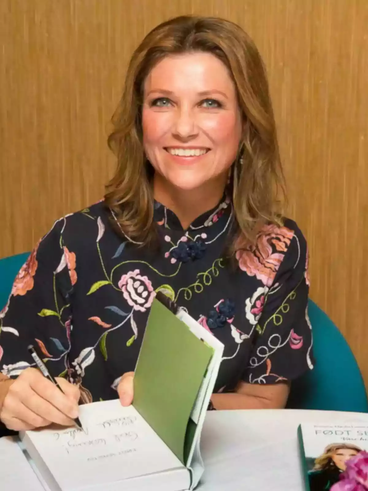 Smiling woman with light brown hair signing a book while sitting at a table, wearing a dark blouse with a floral print Smiling woman with light brown hair signing a book while sitting at a table, wearing a dark blouse with a floral print
