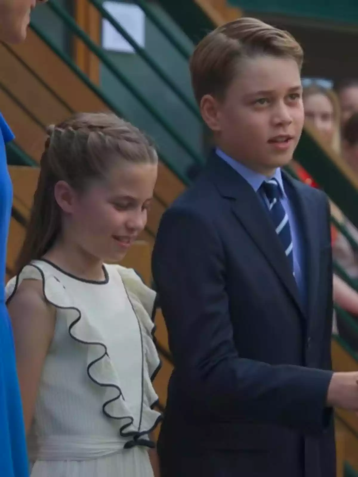 Two well-dressed children, one is Princess Charlotte in a white ruffled dress and the other is Prince George in a blue suit and tie, are standing together at a formal event. Two well-dressed children, one is Princess Charlotte in a white ruffled dress and the other is Prince George in a blue suit and tie, are standing together at a formal event.