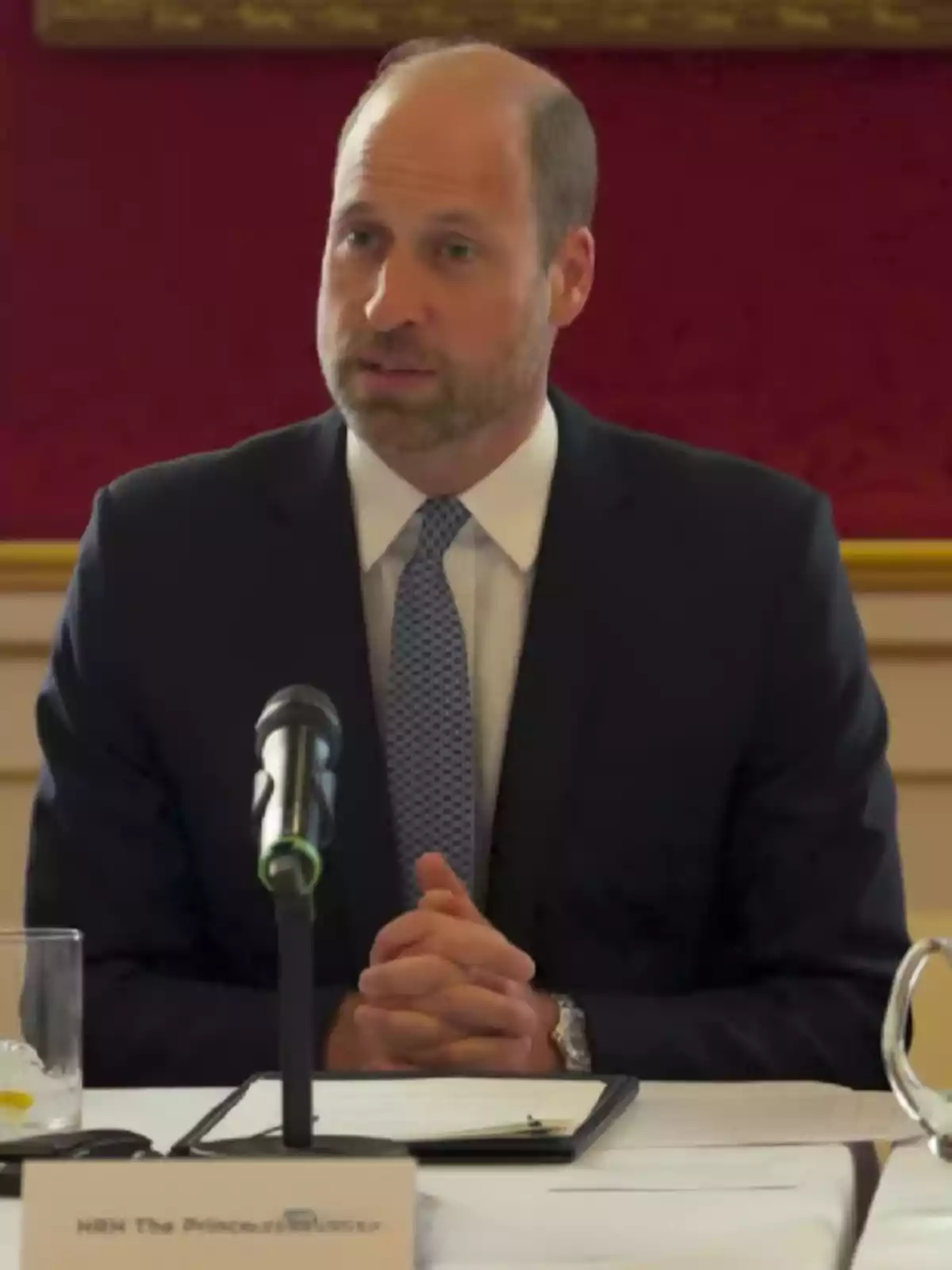 Man in a suit and blue tie speaking in front of a microphone at a conference table Man in a suit and blue tie speaking in front of a microphone at a conference table