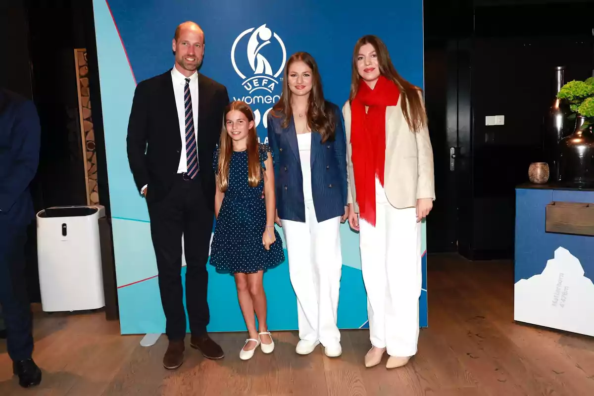 Leonor, Sofía, Prince William, and his daughter Charlotte pose smiling in front of a blue background with the UEFA Women’s Euro logo; two of them are wearing white pants and a red scarf, while a girl is wearing a blue dress with white polka dots. Leonor, Sofía, Prince William, and his daughter Charlotte pose smiling in front of a blue background with the UEFA Women’s Euro logo; two of them are wearing white pants and a red scarf, while a girl is wearing a blue dress with white polka dots.