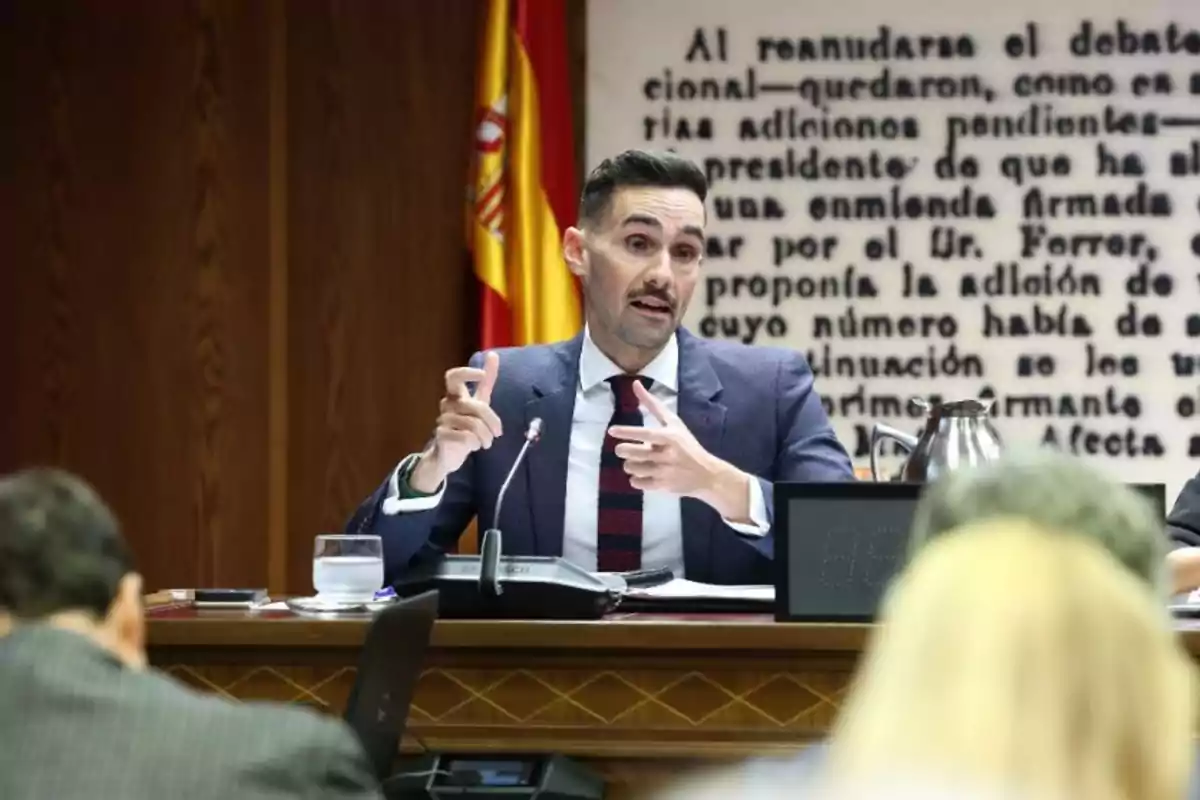 Hombre de traje hablando en una reunión oficial con una bandera de España y un texto grande en la pared de fondo