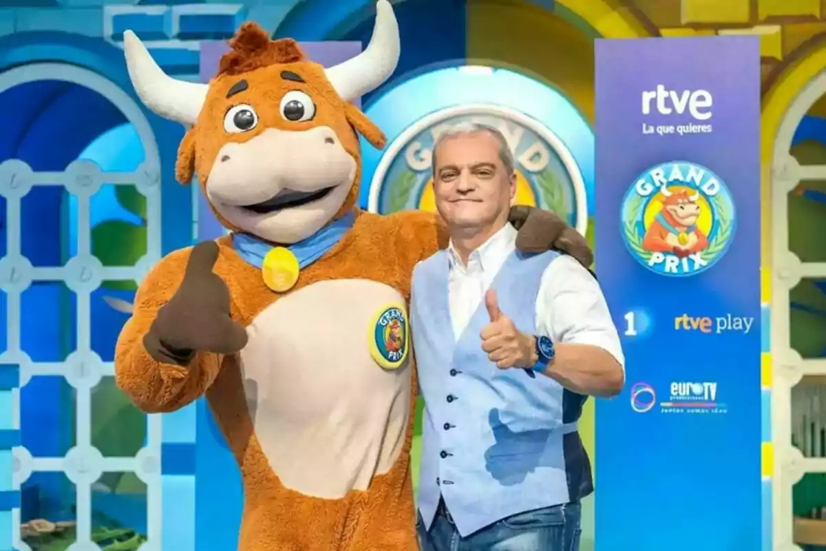 Un hombre sonriente con chaleco azul posa junto a una mascota de toro en un colorido set de televisión del programa Grand Prix Un hombre sonriente con chaleco azul posa junto a una mascota de toro en un colorido set de televisión del programa Grand Prix
