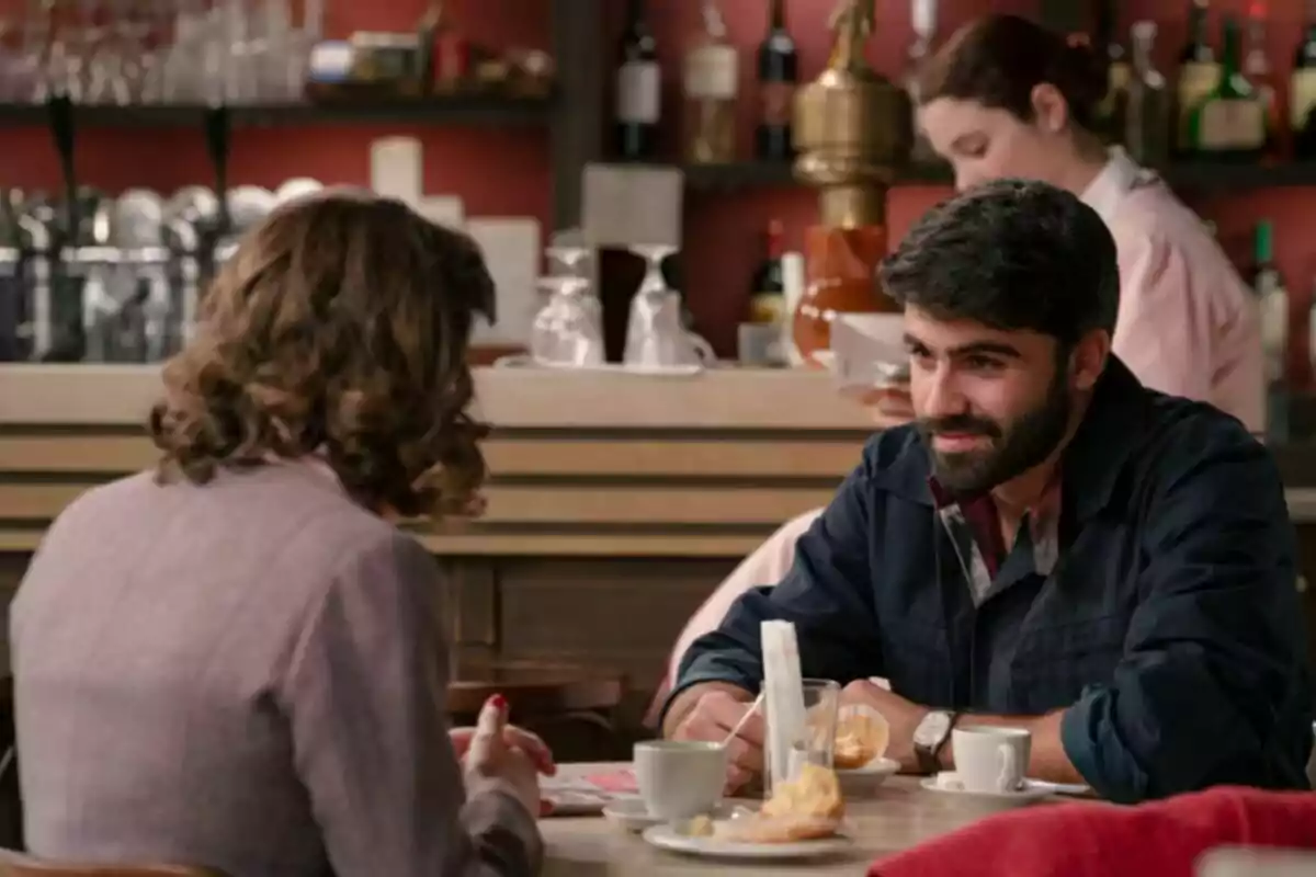 Dos personas conversan en una cafetería mientras disfrutan de café y repostería, al fondo una camarera trabaja detrás de la barra. Se trata de los personajes de Raúl y Claudia en 'Sueños de Libertad'. Dos personas conversan en una cafetería mientras disfrutan de café y repostería, al fondo una camarera trabaja detrás de la barra. Se trata de los personajes de Raúl y Claudia en 'Sueños de Libertad'.