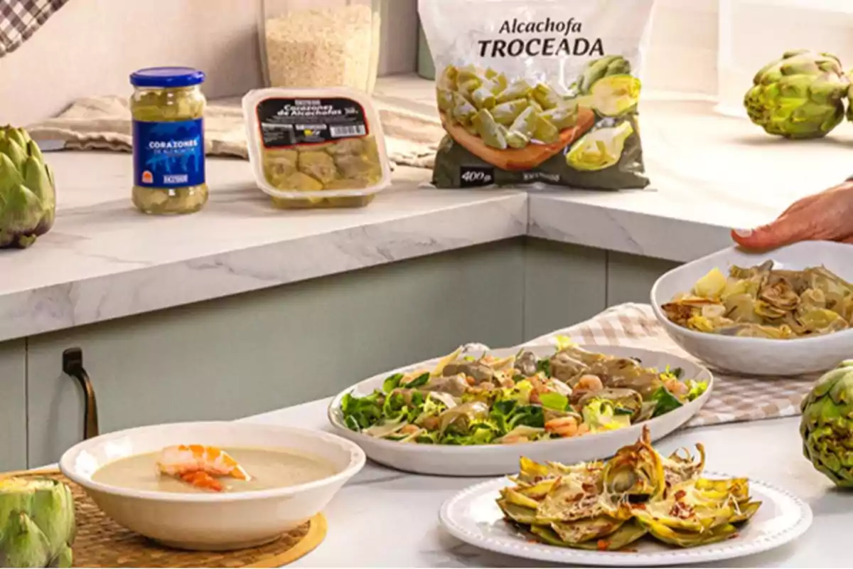 En una cocina se observan varios platos preparados con alcachofa, junto a envases de corazones de alcachofa y una bolsa de alcachofa troceada En una cocina se observan varios platos preparados con alcachofa, junto a envases de corazones de alcachofa y una bolsa de alcachofa troceada