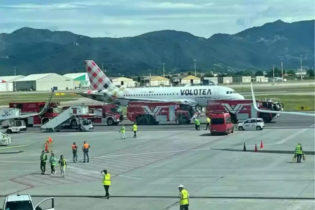 Un avión de la aerolínea Volotea está detenido en la pista rodeado de camiones de bomberos y personal de emergencia mientras varias personas con chalecos de seguridad caminan por el área del aeropuerto con montañas al fondo.